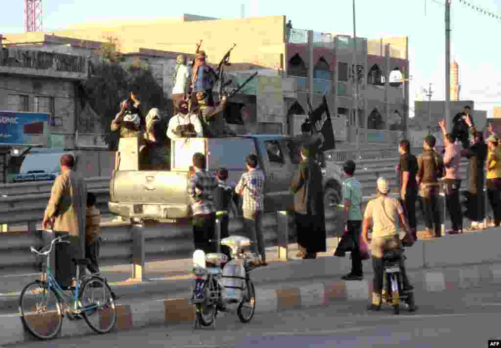 In Fallujah, life goes on. Local Iraqis stand watching an armored vehicle carrying militants holding an Islamic jihadist flag.